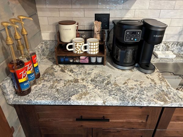 Coffee bar with a dual coffee maker, mugs, and flavored syrups on a granite counter