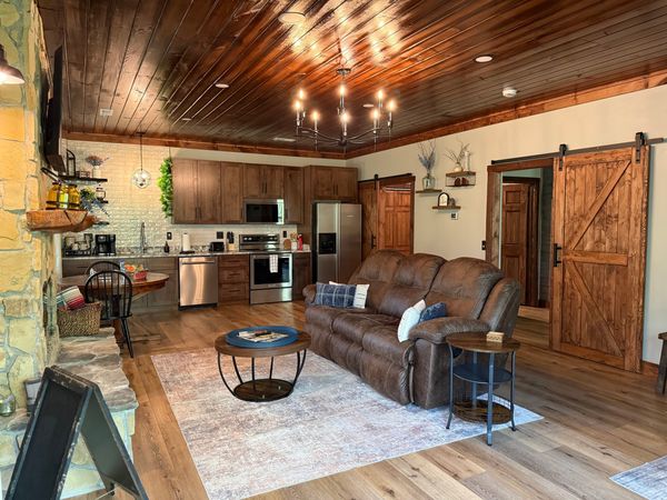 Open living room with a leather reclining sofa and round coffee table, the kitchen behind, under a wood-plank ceiling