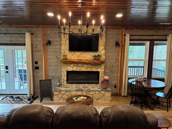 Living room with a stone fireplace, wall-mounted TV, iron chandelier, wood-plank ceiling, and French doors