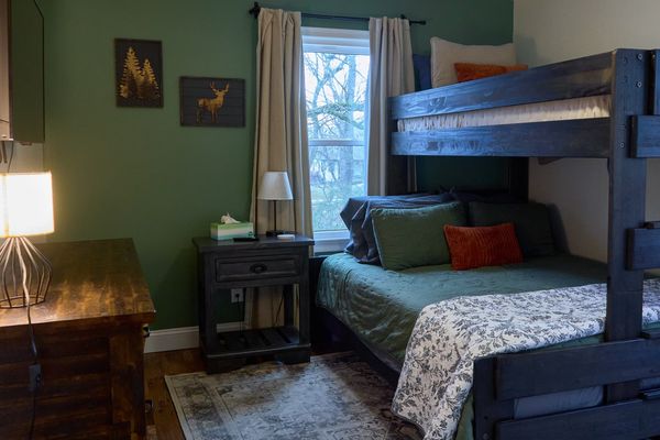 Bunk bed with bedding by a window, with a nightstand and a dresser with a lamp