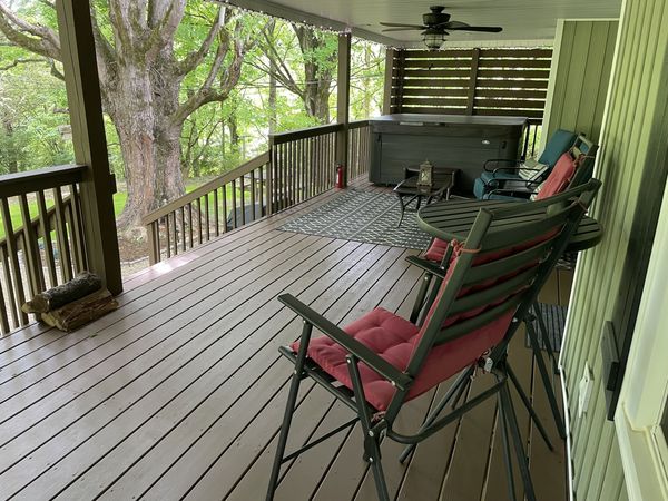 Covered deck with a ceiling fan, cushioned lounge chairs, and the hot tub at the far end under big shade trees