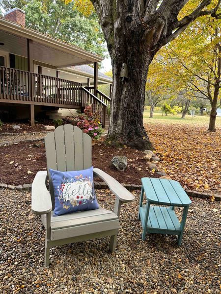 Adirondack chair with a Hello pillow and a side table by the fire pit, with fall leaves and the deck behind