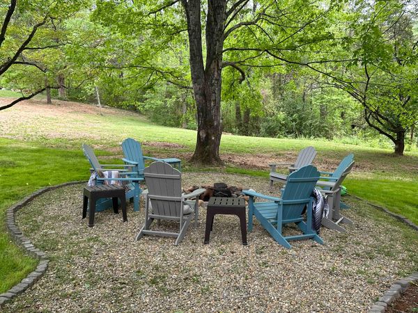 Adirondack chairs around a fire pit on a gravel circle at the edge of the lawn