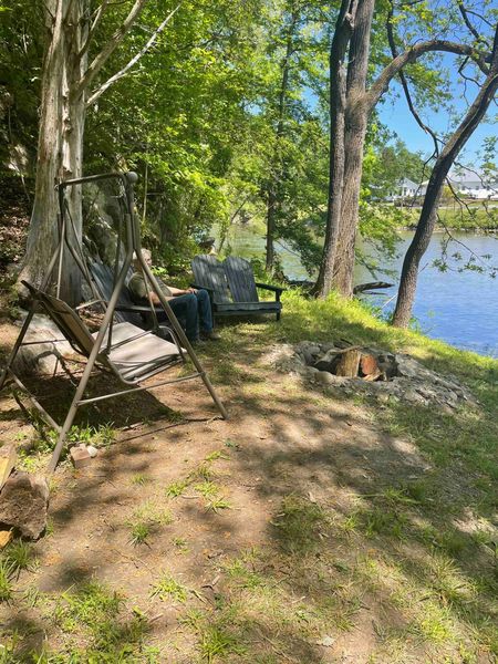 Porch swing, Adirondack chairs, and a stone fire ring on the riverbank, with the river through the trees