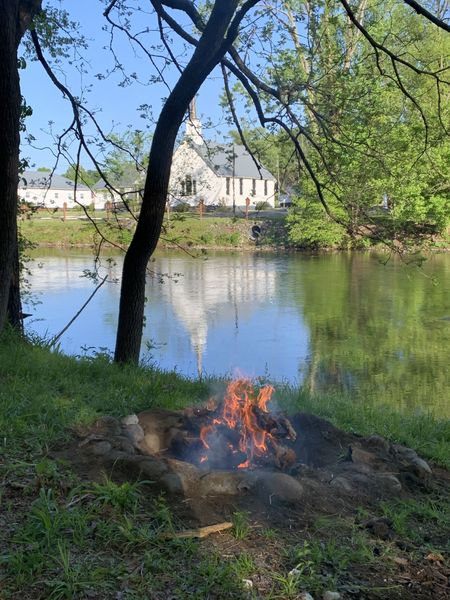 Campfire burning in a stone ring on the riverbank, with a white church reflected across the river