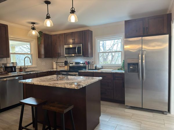 Kitchen with dark wood cabinets, a granite island with two stools, pendant lights, and a stainless refrigerator