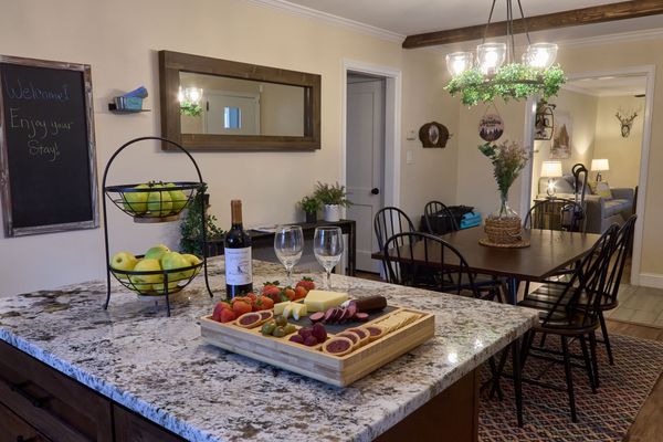 Granite kitchen island set with fruit, a snack board, wine, and glasses, with the dining table beyond