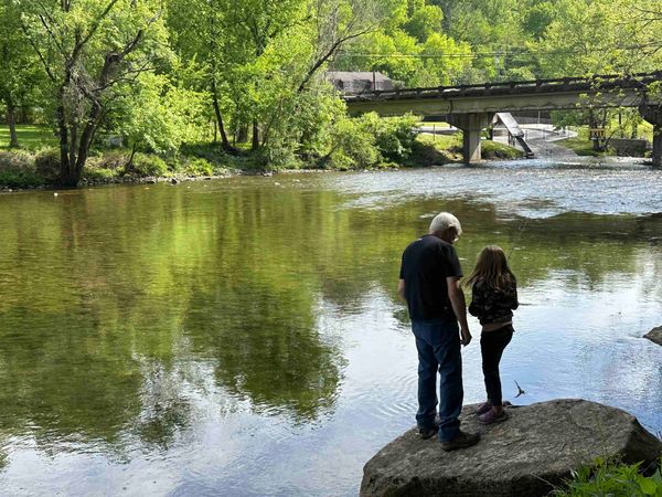 Two people standing on a large rock at the edge of Little River, with a bridge in the distance